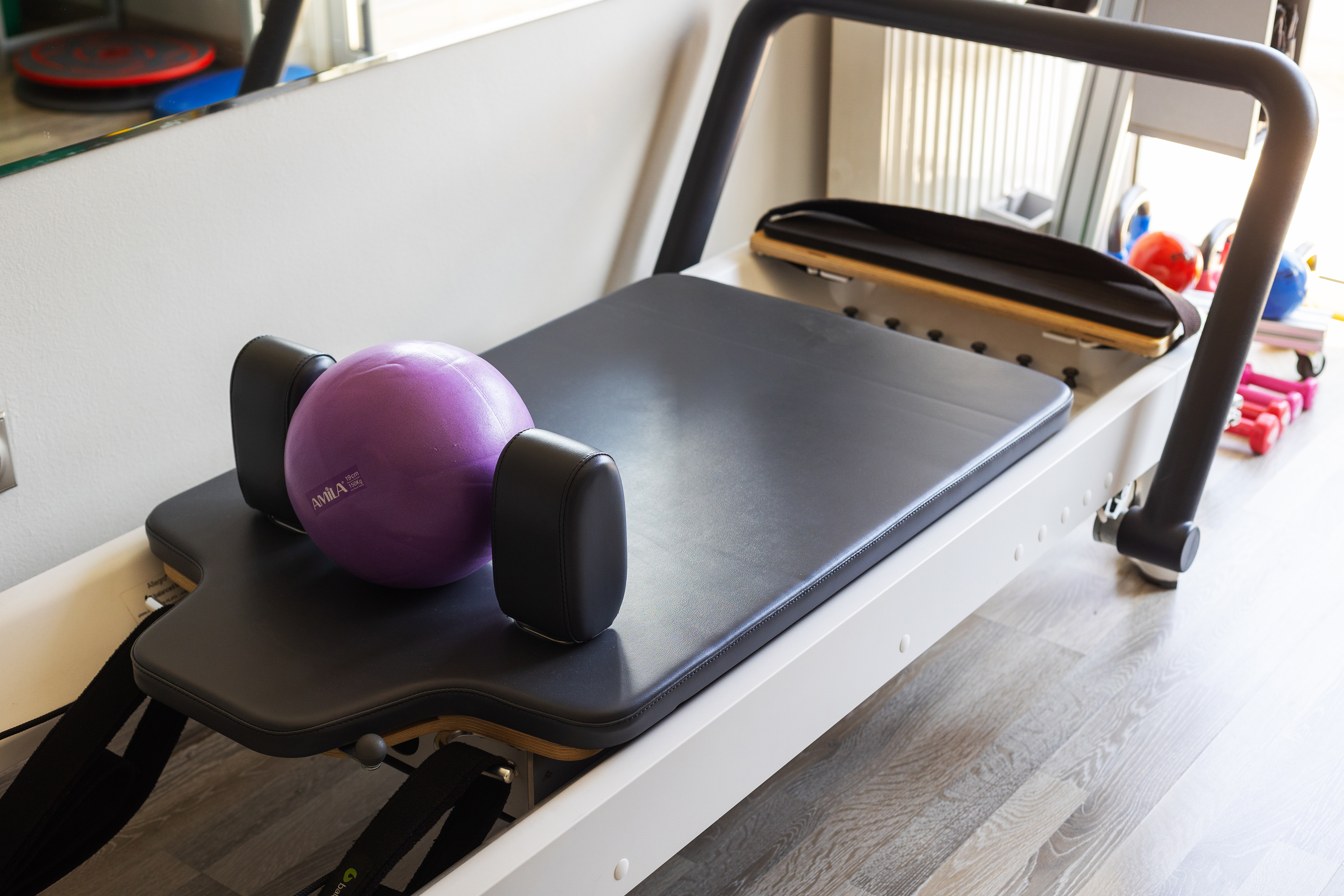 Reformer Pilates με μαύρο στρώμα και μοβ μπάλα άσκησης, δίπλα σε καθρέφτη και παράθυρο.