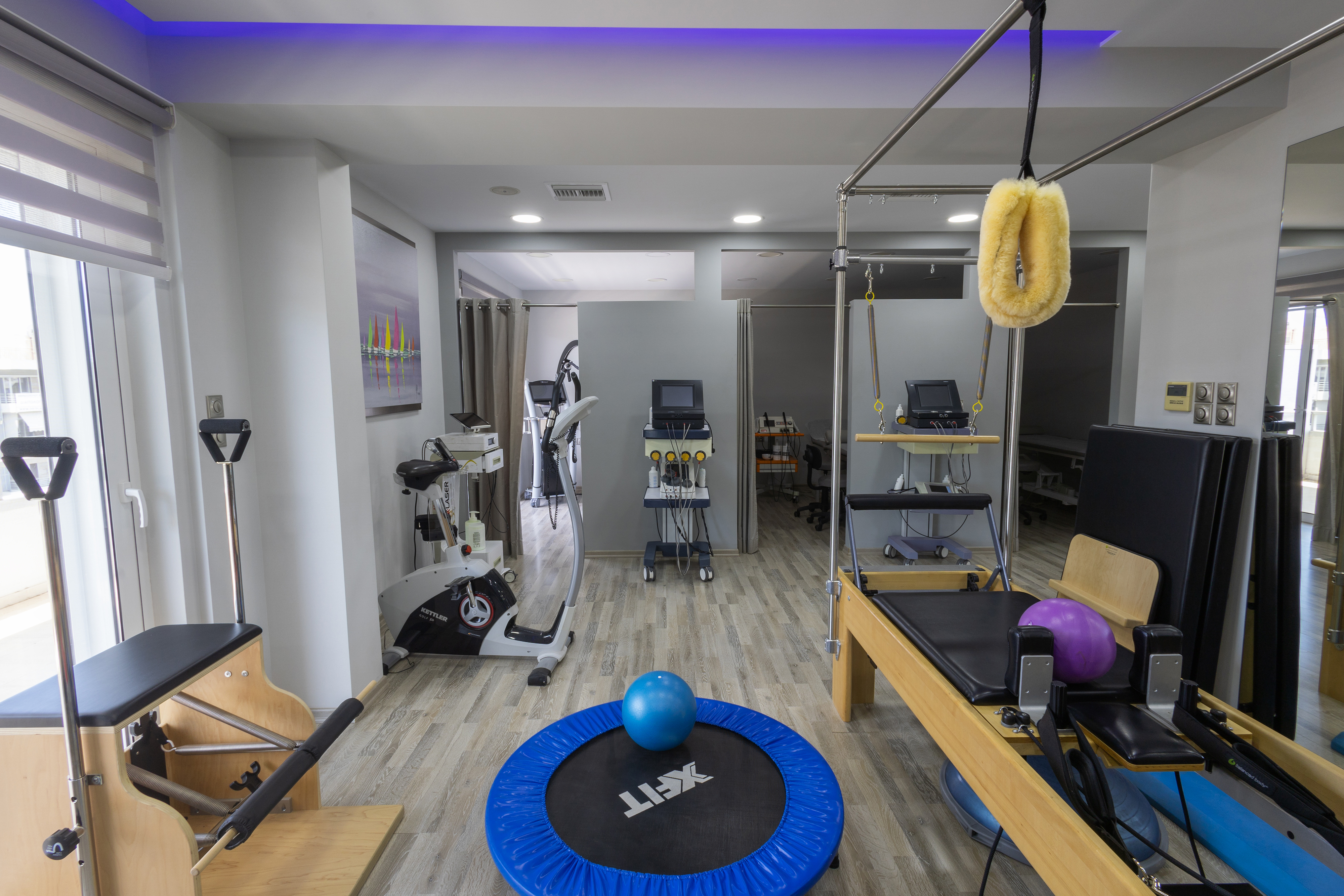 Χώρος φυσικοθεραπείας με trampoline Kettler, συσκευές Pilates και ποδήλατο, φωτισμένος με LED οροφής.