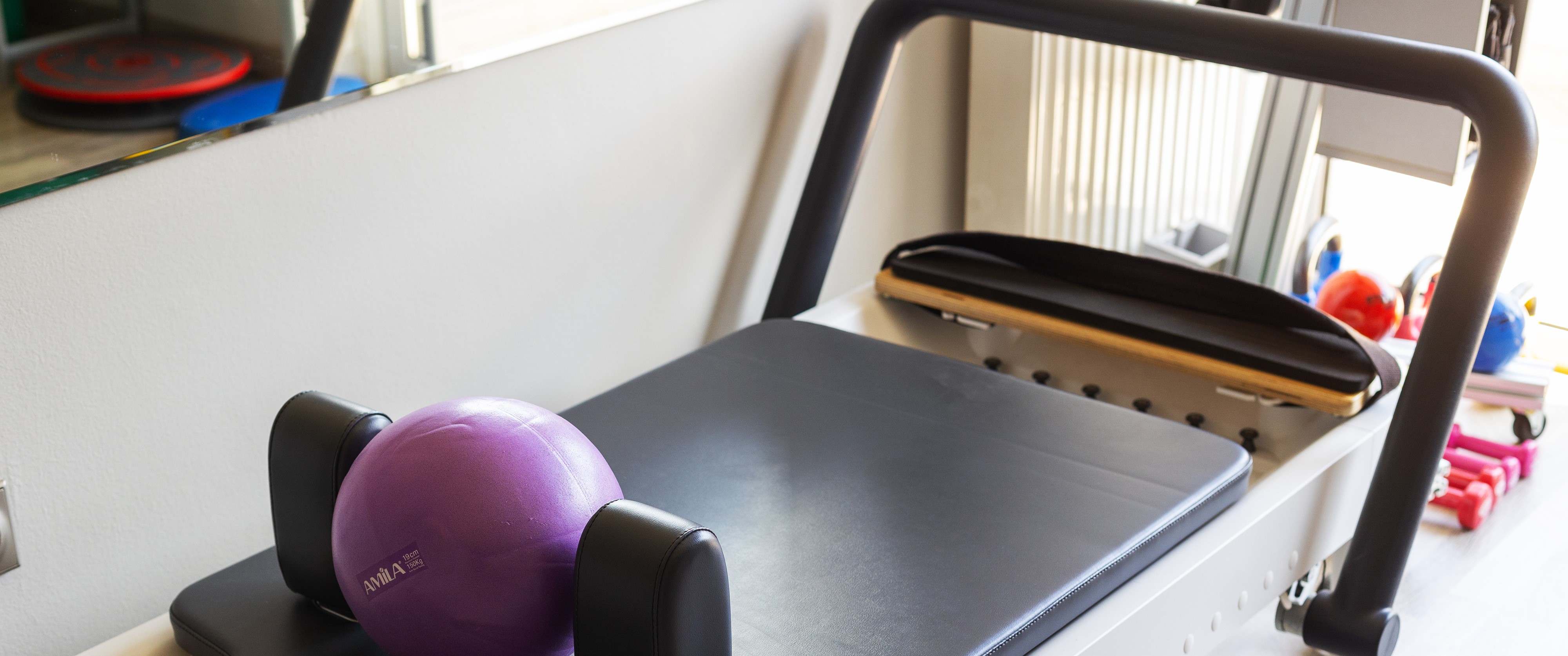 Συσκευή Pilates Reformer με μαύρη επιφάνεια και τοποθετημένη μωβ μπάλα γυμναστικής, δίπλα σε καθρέφτη και βαράκια.