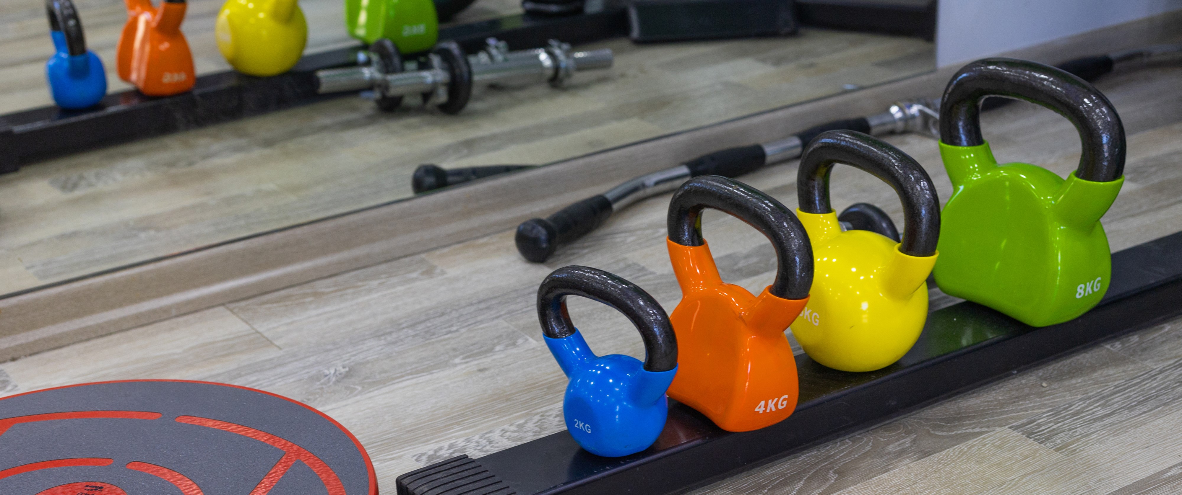 Πολύχρωρα kettlebells από 2 έως 8 κιλά τοποθετημένα μπροστά σε καθρέφτη, μαζί με βάρη και μπάρες ασκήσεων.